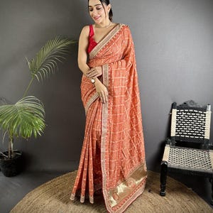 May include: An orange saree with a grid pattern and gold trim. The saree is draped over a person wearing a red top. The saree has intricate embroidery along the borders and a decorative gold band at the hem. A traditional Indian garment.