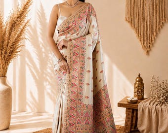 May include: A cream-colored saree featuring an intricate border with pink, green, and gold paisley patterns. The fabric has a smooth, satin-like finish with small floral motifs scattered across the body, finished with decorative tassels at the end.