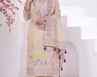 May include: A beige salwar kameez ensemble featuring a tunic, pants, and a matching scarf. The outfit is adorned with floral patterns in shades of purple and green. The scarf has purple tassels. The outfit is worn with brown flats.