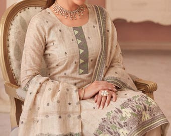 May include: A beige salwar kameez set. The long-sleeved tunic features a round neckline with a green and gray geometric design. The matching scarf and pants have a floral border. The woman is wearing a necklace and ring.