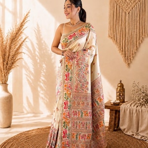 May include: A cream-colored saree featuring intricate, multi-colored floral and animal motifs in shades of red, green, orange, and blue. The garment includes a wide, detailed border with repeating patterns and a fringed hem at the bottom edge.