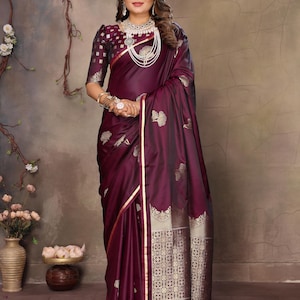 May include: A formal burgundy saree with gold detailing and floral patterns. The saree has a detailed border and a matching blouse with patterned sleeves. The outfit is accessorised with jewellery.
