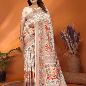 May include: Ivory-colored sari with intricate floral and figurative patterns in red, orange, green, and blue. The sari features a detailed border and is draped over a person. The outfit includes a matching blouse and gold jewelry.