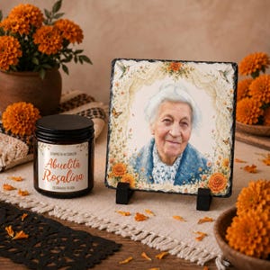 May include: A square slate plaque featuring a portrait of an elderly person framed by orange marigolds, lace, and butterflies, displayed on a black stand next to a candle jar labeled "Siempre en mi corazón Abuelita Rosalina Celebrando tu vida."