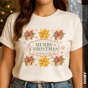 May include: Ivory-colored t-shirt with a festive "Merry Christmas" design. The design features poinsettias in red, orange, and yellow, along with small hearts and sewing-themed elements. The text "Threads of Joy and Love" is also included.