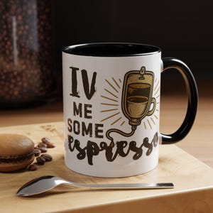 May include: White ceramic coffee mug with black interior and handle. The mug features the text "IV ME SOME espresso" in brown, along with a graphic of a coffee drip bag. A spoon and a macaron are in the foreground.