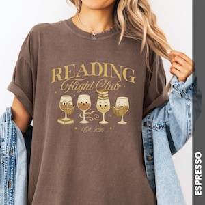 Puede incluir: Camiseta color espresso con la frase "READING Flight Club" en letras doradas. Debajo, cuatro copas de vino de dibujos animados, cada una con una expresi&oacute;n diferente y un libro. La camiseta es de estilo c&oacute;modo e informal.