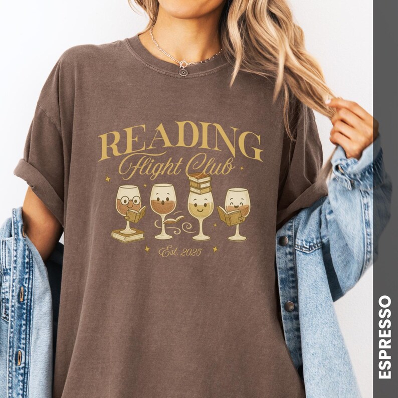 Puede incluir: Camiseta color espresso con la frase "READING Flight Club" en letras doradas. Debajo, cuatro copas de vino de dibujos animados, cada una con una expresi&oacute;n diferente y un libro. La camiseta es de estilo c&oacute;modo e informal.