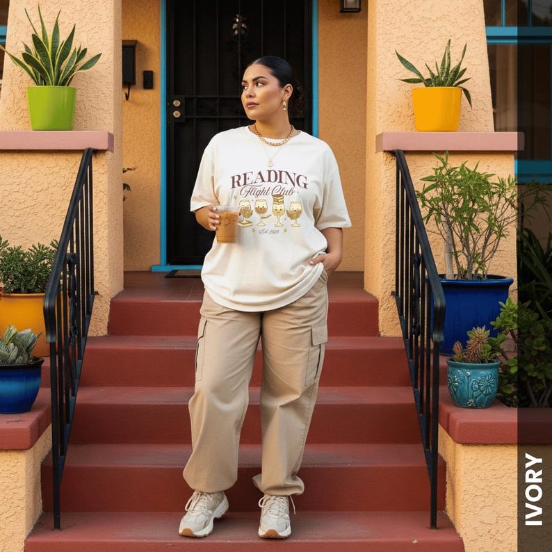 Puede incluir: Una persona con una camiseta color crema con el texto "Reading Flight Club" y un gr&aacute;fico de bebidas de caf&eacute;. La persona tambi&eacute;n lleva pantalones cargo color caqui y zapatillas blancas. El fondo incluye una escalera roja y plantas en macetas.