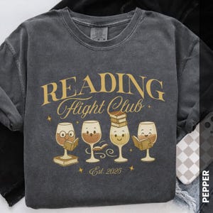 Puede incluir: Sudadera gris oscuro con el texto "READING Flight Club" en letras doradas. Debajo del texto hay cuatro copas de vino de dibujos animados, cada una con una expresi&oacute;n diferente y sosteniendo o apilando libros. El texto "Est. 2025" est&aacute; debajo.