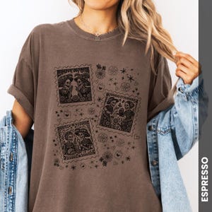 May include: Espresso-colored t-shirt featuring three postage stamp-style illustrations of skeleton figures in festive attire. The stamps are surrounded by floral and heart motifs. The shirt has a relaxed fit and rolled-up sleeves.
