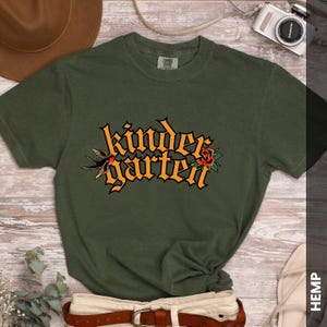 Puede incluir: Camiseta verde oliva con la palabra "kindergarten" en fuente gótica naranja, acompañada de una golondrina y una rosa roja. La camiseta se combina con un cinturón marrón y una cámara.