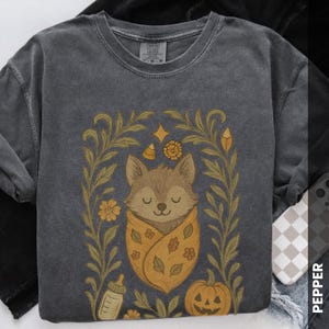 May include: Dark gray sweatshirt featuring a cartoon wolf wrapped in a yellow blanket with fall-themed illustrations. The design includes a pumpkin, flowers, and a baby bottle, framed by a floral border. The sweatshirt has a vintage, washed look.