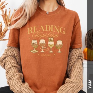 Puede incluir: Camiseta color &oacute;xido con el texto "READING Flight Club" en letras doradas. Debajo, cuatro copas de vino de dibujos animados, cada una con un libro. La camiseta tambi&eacute;n tiene "Est. 2025" impreso.