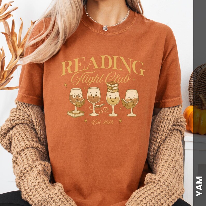 Puede incluir: Camiseta color &oacute;xido con el texto "READING Flight Club" en letras doradas. Debajo, cuatro copas de vino de dibujos animados, cada una con un libro. La camiseta tambi&eacute;n tiene "Est. 2025" impreso.