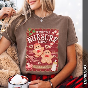 May include: Espresso-colored t-shirt featuring a festive design. The graphic includes the text "North Pole Nursery Lounge" with gingerbread people, a candy cane, and a baby bottle. The shirt also has the text "Milk, Cocoa & Cuddles" and "Swaddled with Love."