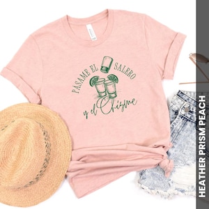 Könnte beinhalten: Ein pfirsichfarbenes T-Shirt mit hochgekrempelten Ärmeln. Das Shirt zeigt eine grüne Grafik von zwei Schnapsgläsern mit Limettenspalten und dem Text "Pásame el Salero y el Chisme". Ein Strohhut und Jeansshorts sind ebenfalls zu sehen.