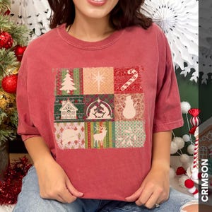 May include: A red-toned t-shirt featuring a patchwork design with Christmas-themed images. The design includes a Christmas tree, star, candy cane, house, nativity scene, snowman, pig, deer, and snowflakes. The word "CRIMSON" is printed vertically on the right side.