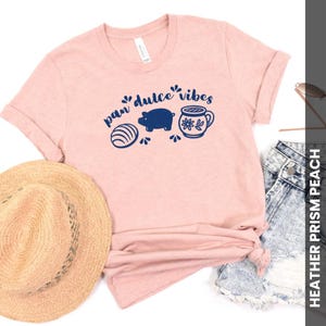 May include: A light pink t-shirt with rolled-up sleeves featuring a navy blue graphic that reads "pan dulce vibes." The graphic includes a sweet bread, a pig, and a decorated mug. A straw hat and denim shorts are also visible.