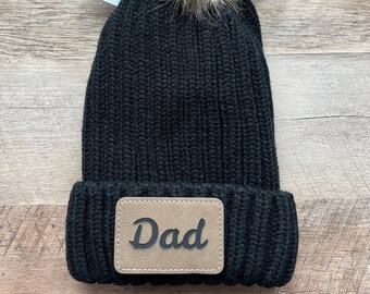 May include: Black knit beanie with a brown faux fur pom pom and a brown leather patch that says "Dad".