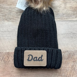 May include: Black knit beanie with a brown faux fur pom pom and a brown leather patch that says "Dad".