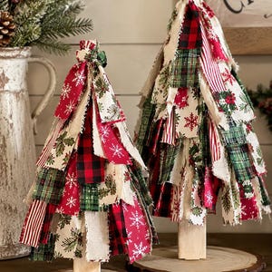 May include: Two decorative fabric trees constructed from layered strips of cloth in red, green, white, and black patterns, including snowflakes, plaid, and holly prints. Each tree stands on a light-colored wooden post and a circular wood slice base.
