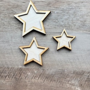 May include: Three wooden stars of different sizes, all with a white center and a light brown border. The stars are arranged in a staggered pattern on a light brown wooden background.