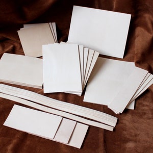 May include: A set of white craft paper in various sizes and shapes, including squares, rectangles, and strips. The paper is arranged on a brown fabric surface.