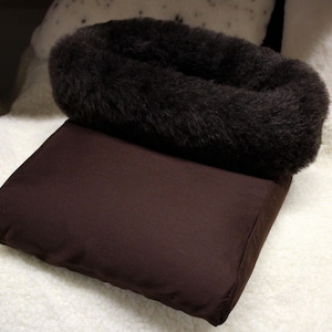 May include: A brown rectangular cushion with a dark brown, fluffy, fur-like trim. The cushion is on a white, textured surface. The cushion appears to be made of a soft, plush material.