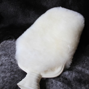 Może przedstawiać: Biała butelka z ciepłą wodą z imitacji futra, z białą plastikową podstawą i uchwytem. Butelka z ciepłą wodą leży na ciemnoszarym tle.