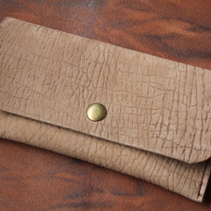 Könnte beinhalten: Eine hellbraune Lederbörse mit strukturierter Oberfläche und einem einzigen Messingknopfverschluss.