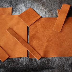 May include: Assortment of brown leather pieces in various shapes and sizes, including squares, rectangles, and strips. The leather is a light brown color and has a textured surface.