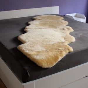 Pode incluir: Um tapete de pele de ovelha bege claro está colocado sobre uma cama cinza escuro. O tapete tem uma forma natural e irregular e uma aparência macia e texturizada. A cama é branca com um lençol cinza escuro. O fundo inclui uma parede roxa e uma mesa de cabeceira branca.