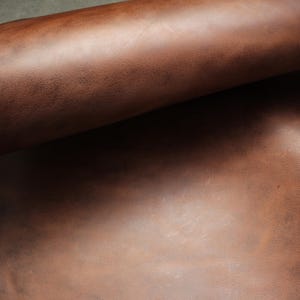 May include: Close-up of a rolled-up sheet of brown leather. The leather has a smooth, textured surface with variations in color, ranging from light to dark brown. The image highlights the natural grain and texture of the material.