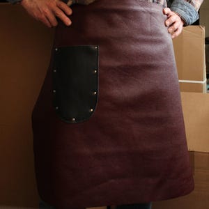 Può includere: Un grembiule in pelle bordeaux con una tasca in pelle nera e rivetti argentati. Il grembiule è indossato da una persona, ma la persona non è visibile nell'immagine.
