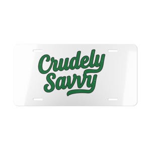 May include: A rectangular white license plate with four mounting slots, featuring the text "Crudely Savvy" written in a bold, dark green, cursive script font. The lettering has a slightly distressed, textured appearance across the surface.