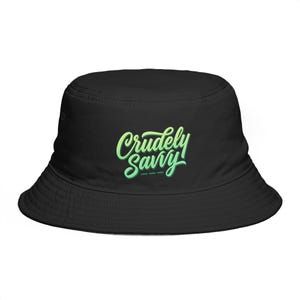 May include: A black bucket hat featuring the text Crudely Savvy printed in a light green, cursive script font on the front center. The hat has a rounded crown and a downward-sloping brim, maintaining a solid black color throughout the design.