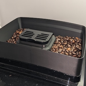 Op de afbeelding: Een zwarte koffiemolenbak gevuld met geroosterde koffiebonen. Een zwart rechthoekig deksel met sleuven ligt bovenop de bonen. De molen staat op een zwart oppervlak.