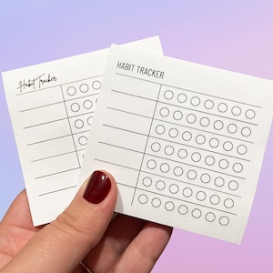 May include: Two white square paper habit tracker cards with black ink. Each card features the text HABIT TRACKER at the top, horizontal lines for writing, and a vertical column of empty circles for marking progress on daily tasks.