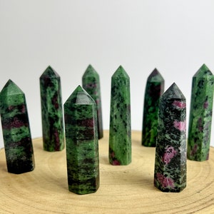 May include: Eight ruby zoisite crystal towers are displayed. The crystals are green and black with red flecks. They are arranged on a wooden surface against a white backdrop. Each tower has a pointed apex.