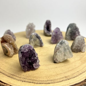 May include: A collection of small amethyst and clear quartz crystal clusters on a wooden surface. The crystals are various shades of purple and clear, and some have a natural, rough texture.