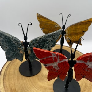 May include: Gemstone butterfly sculptures in green, orange, and red. Each butterfly features detailed wing patterns and is mounted on a black metal stand. The butterflies are displayed on a wooden surface, creating a natural aesthetic.