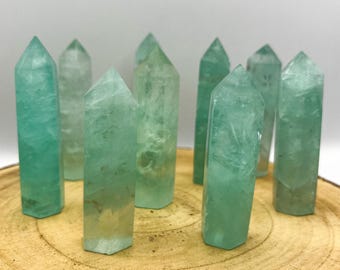 May include: A collection of nine green fluorite crystal points. The crystals vary in shades of green and are arranged on a wooden surface. Each crystal has a pointed top and a rectangular base. The crystals are translucent.