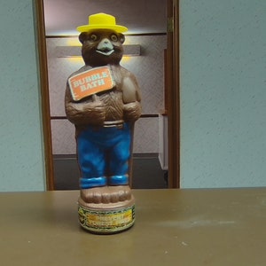 以下が含まれることがあります： 黄色の帽子をかぶって「バブルバス」と書かれた看板を持っている茶色のクマの置物。置物は茶色のクマのデザインと青いジーンズのプラスチックボトルです。ボトルは茶色の表面の上にあります。