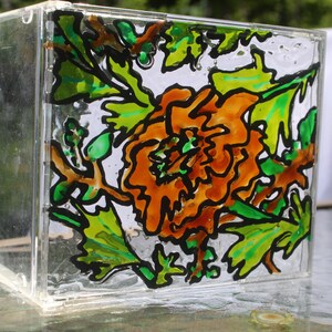 Puede incluir: Una caja de vidrio transparente y cuadrada con un diseño floral pintado a mano. La obra de arte presenta una flor naranja con hojas verdes y tallos marrones, delineados en negro. La caja está sobre una superficie de vidrio.