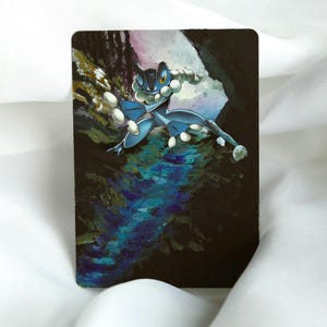 May include: A rectangular card featuring a blue and white cartoon character in a dynamic pose against a dark, textured background. The artwork depicts a creature with large eyes and bulbous features, set in a stylized environment with water and rock formations.