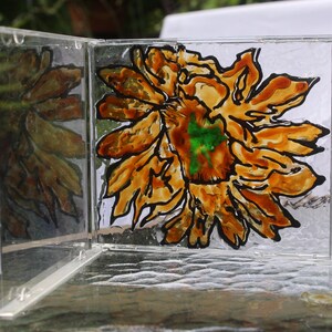 Puede incluir: Una caja acrílica transparente con bisagras que presenta un diseño de girasol pintado a mano. La flor tiene pétalos naranjas y amarillos con un centro verde, delineados en negro. La caja se exhibe sobre una mesa de vidrio, con un fondo verde borroso.
