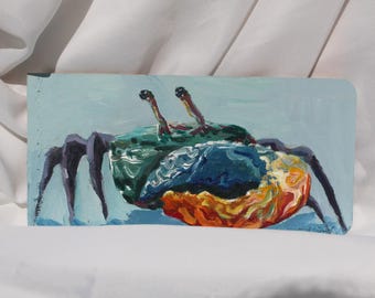 May include: A rectangular painting of a crab. The crab has a green shell, blue and orange body, and dark purple legs. The background is a light blue color. The painting is on a white surface.