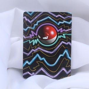 Può includere: Una carta rettangolare con una sfera rossa e bianca con una faccia, circondata da disegni di fulmini colorati su sfondo nero. La carta è posta su uno sfondo di tessuto bianco.