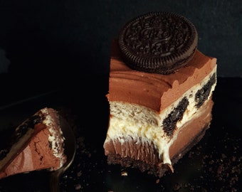 May include: A slice of chocolate cheesecake with a layer of vanilla cheesecake and a chocolate cookie crust, topped with a chocolate cookie. The cheesecake is on a black plate with crumbs.